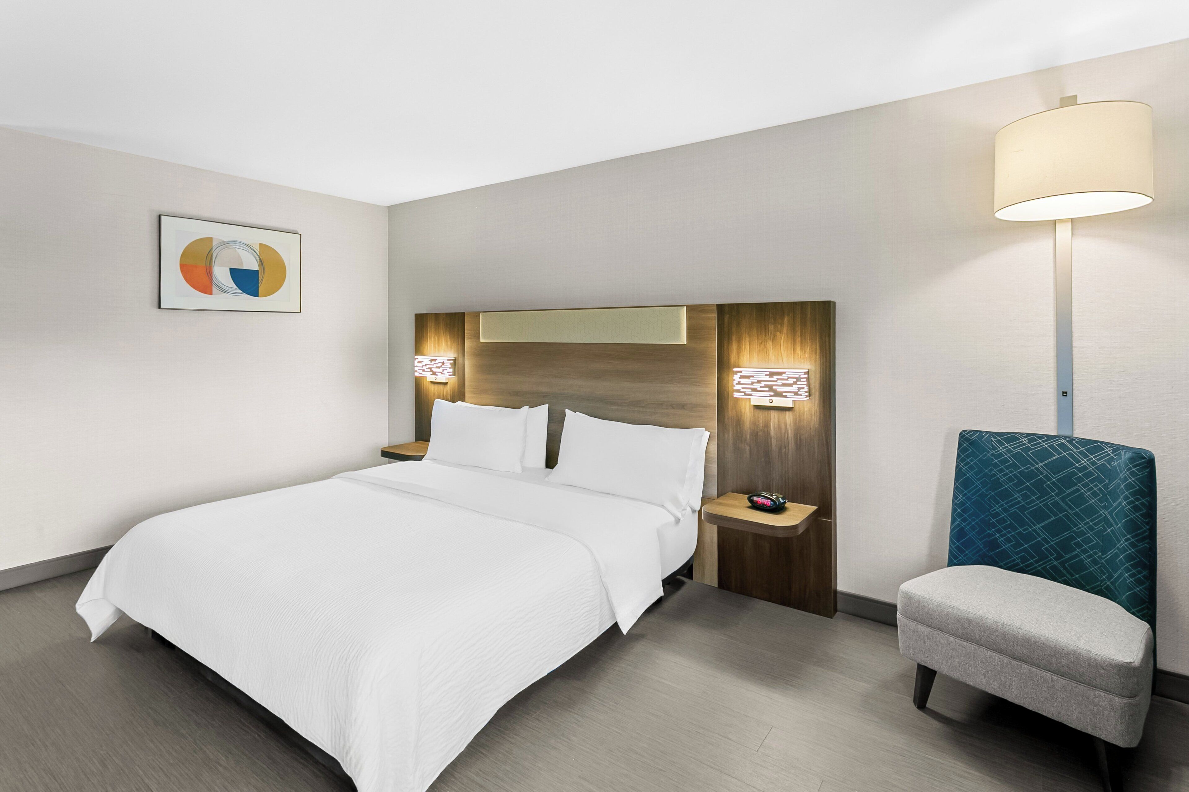 A hotel room with a large bed, a bedside table with a lamp, a chair, and a wall-mounted artwork.