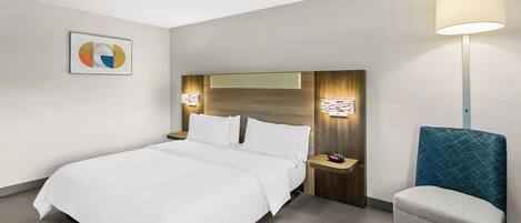 A hotel room with a large bed, a bedside table with a lamp, a chair, and a wall-mounted artwork.
