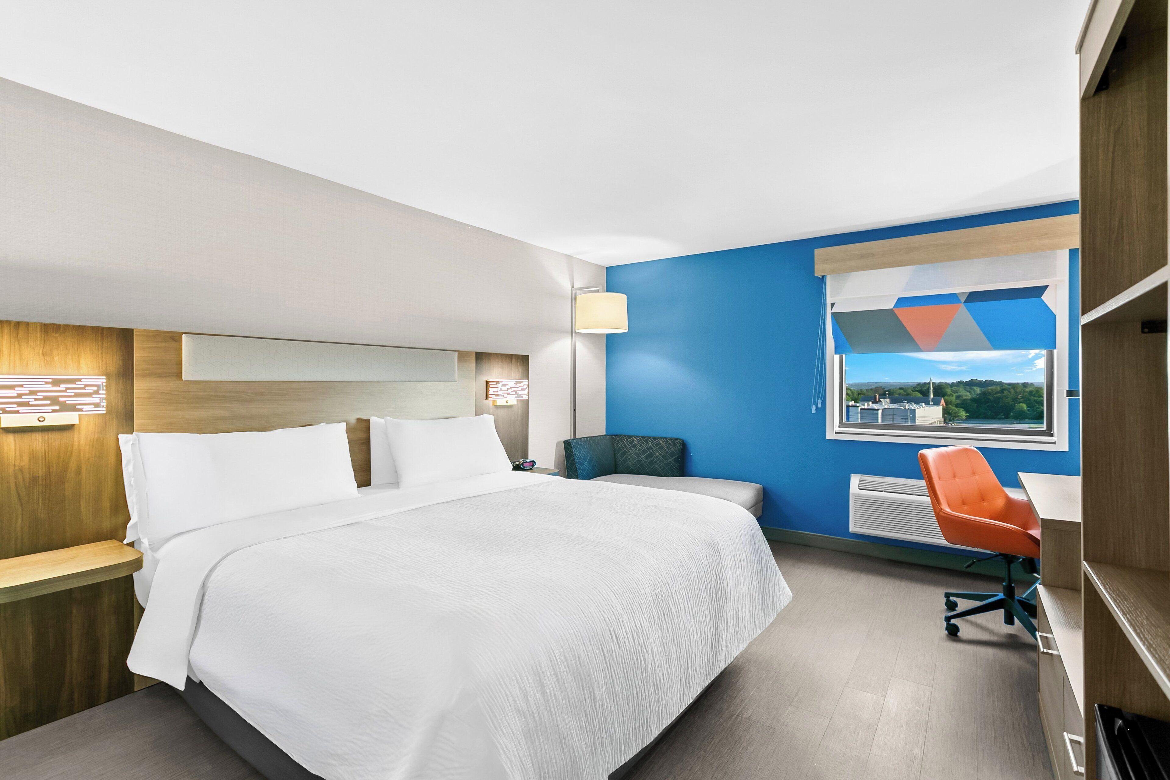 A hotel room with a bed, a desk, a chair, a window with a view, and a lamp.