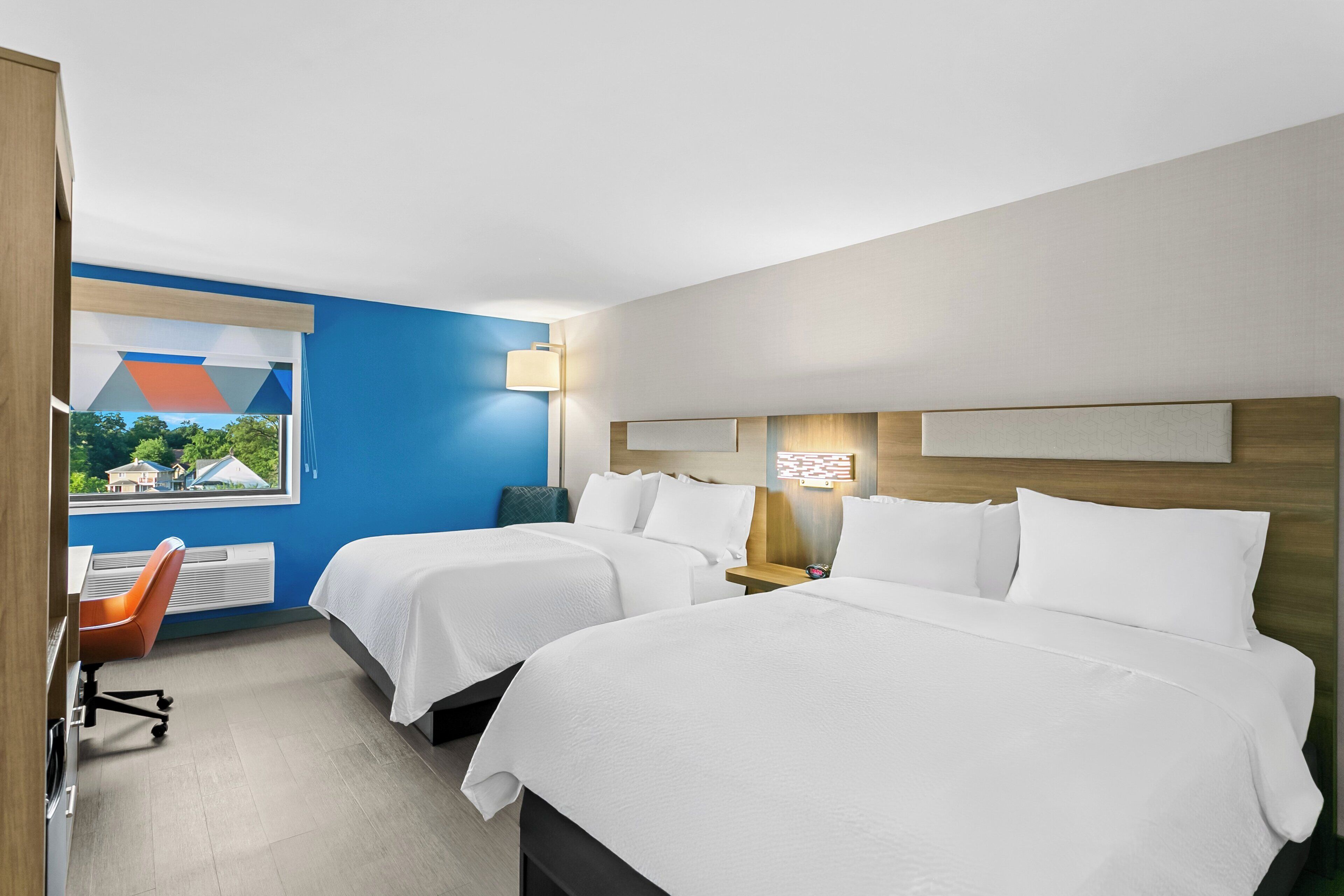 Habitación de hotel con dos camas, un escritorio, una silla y ventana con vista a casas.