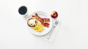 Un plato con huevos revueltos, tocino, un muffin con crema y una manzana sobre una mesa blanca.