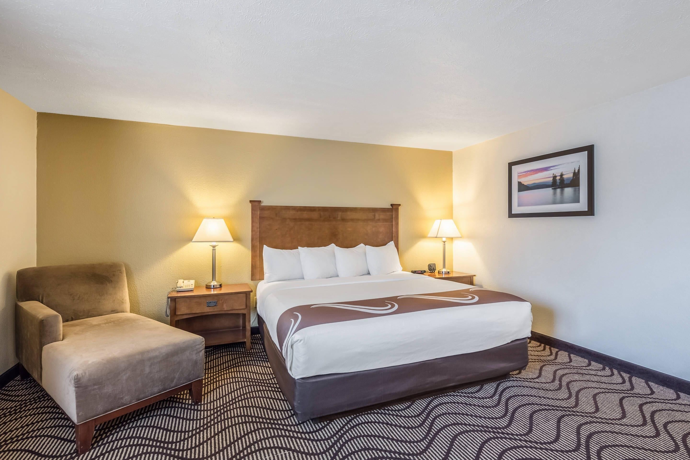 Une chambre d’hôtel avec un lit, deux lampes de chevet, une chaise, une table de chevet et un tableau encadré au mur.
