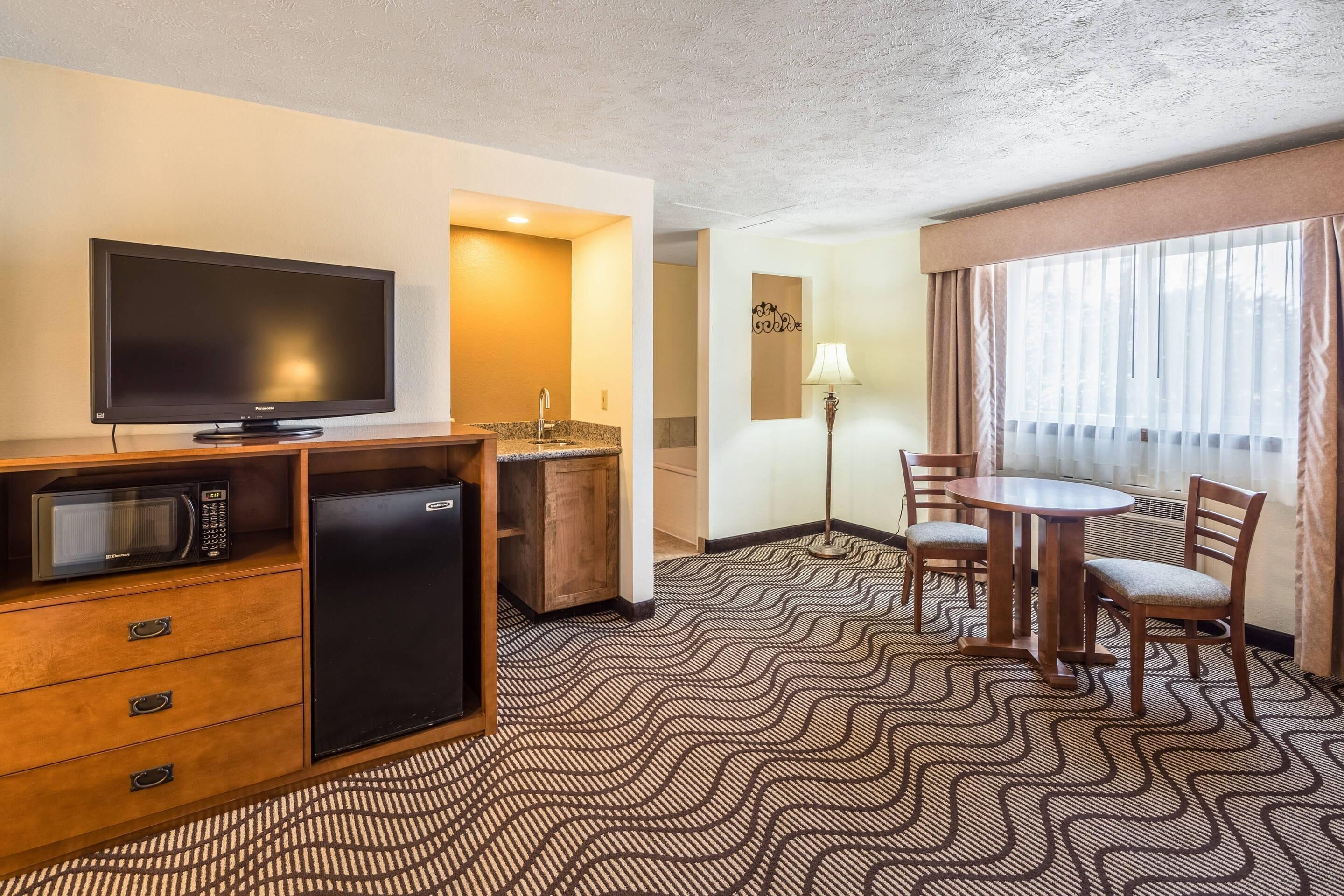 A hotel room with a flat-screen TV, a microwave, a mini-fridge, a dining table with chairs, and a patterned carpet.