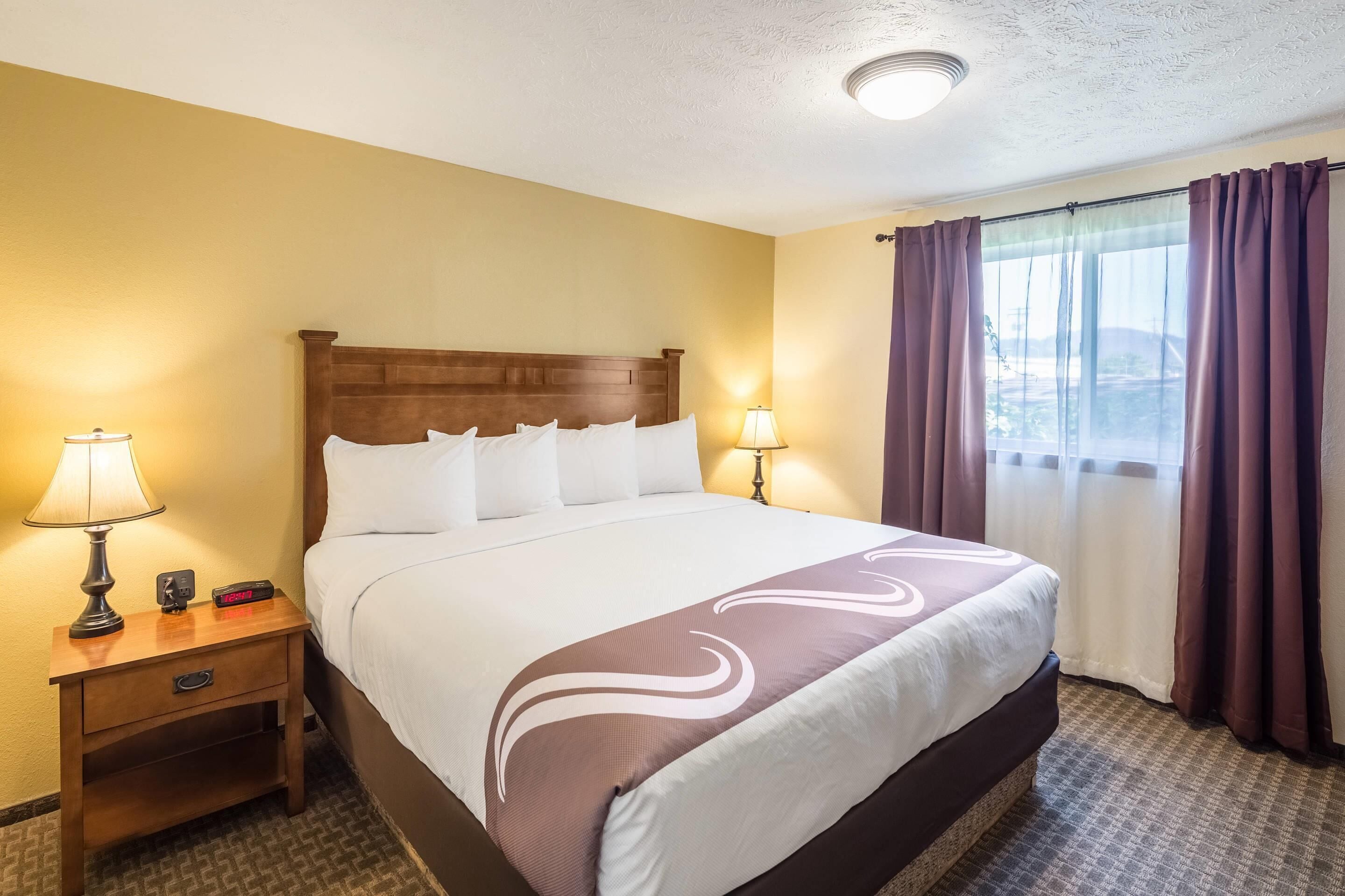 A hotel room with a large bed, two bedside lamps, a nightstand with a lamp, a digital clock, and a window with curtains.