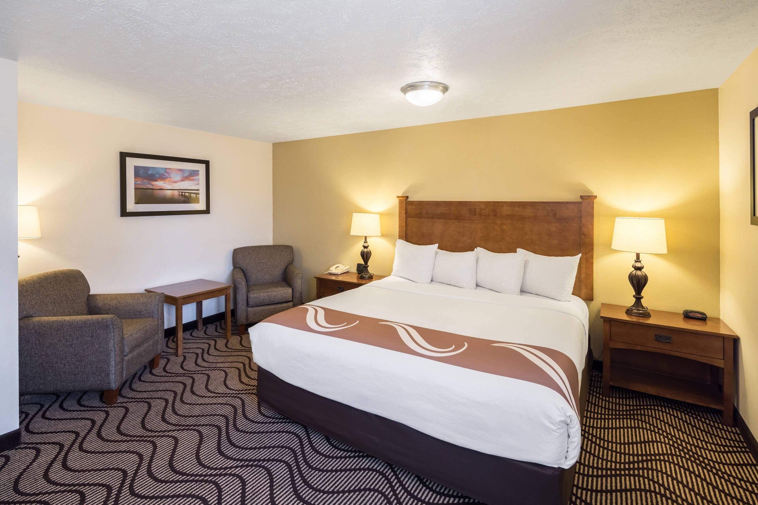 A hotel room with a large bed, two bedside tables, a chair, a small table, and a lamp.