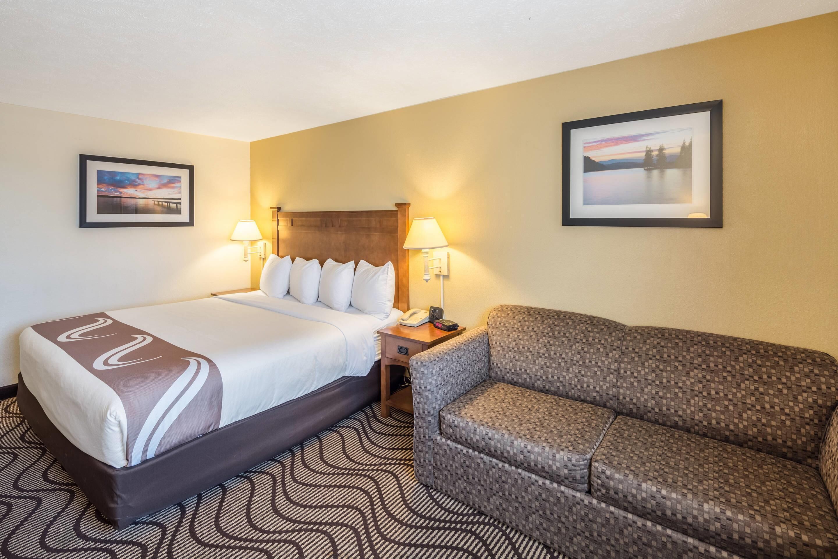 A hotel room with a bed, a sofa, two wall-mounted lamps, and two framed pictures.