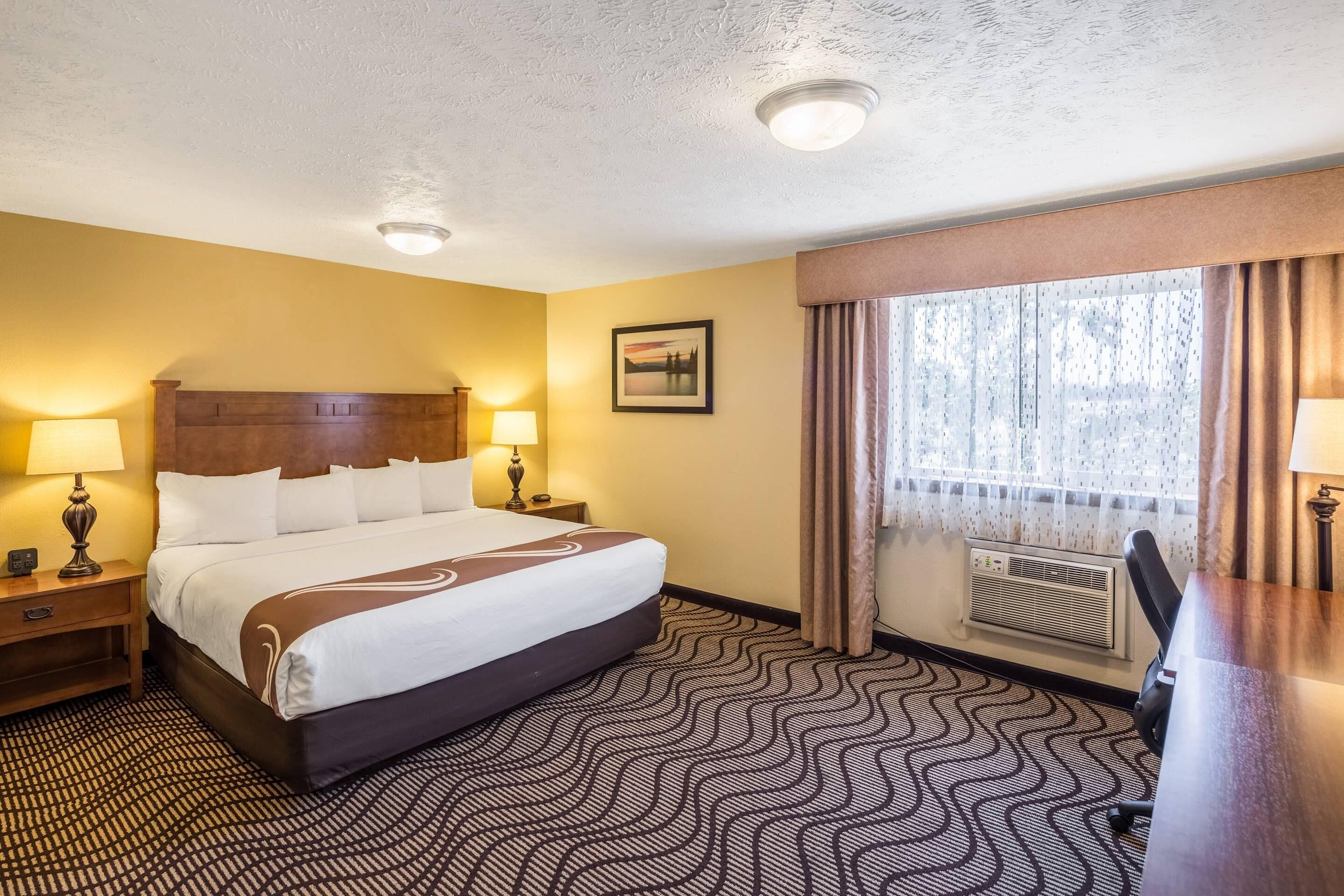 A hotel room with a bed, two bedside lamps, a picture on the wall, a window with curtains, and a desk with a chair.