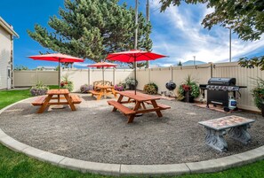Un jardin avec des tables de pique-nique, des parasols rouges, un barbecue et une table décorative.
