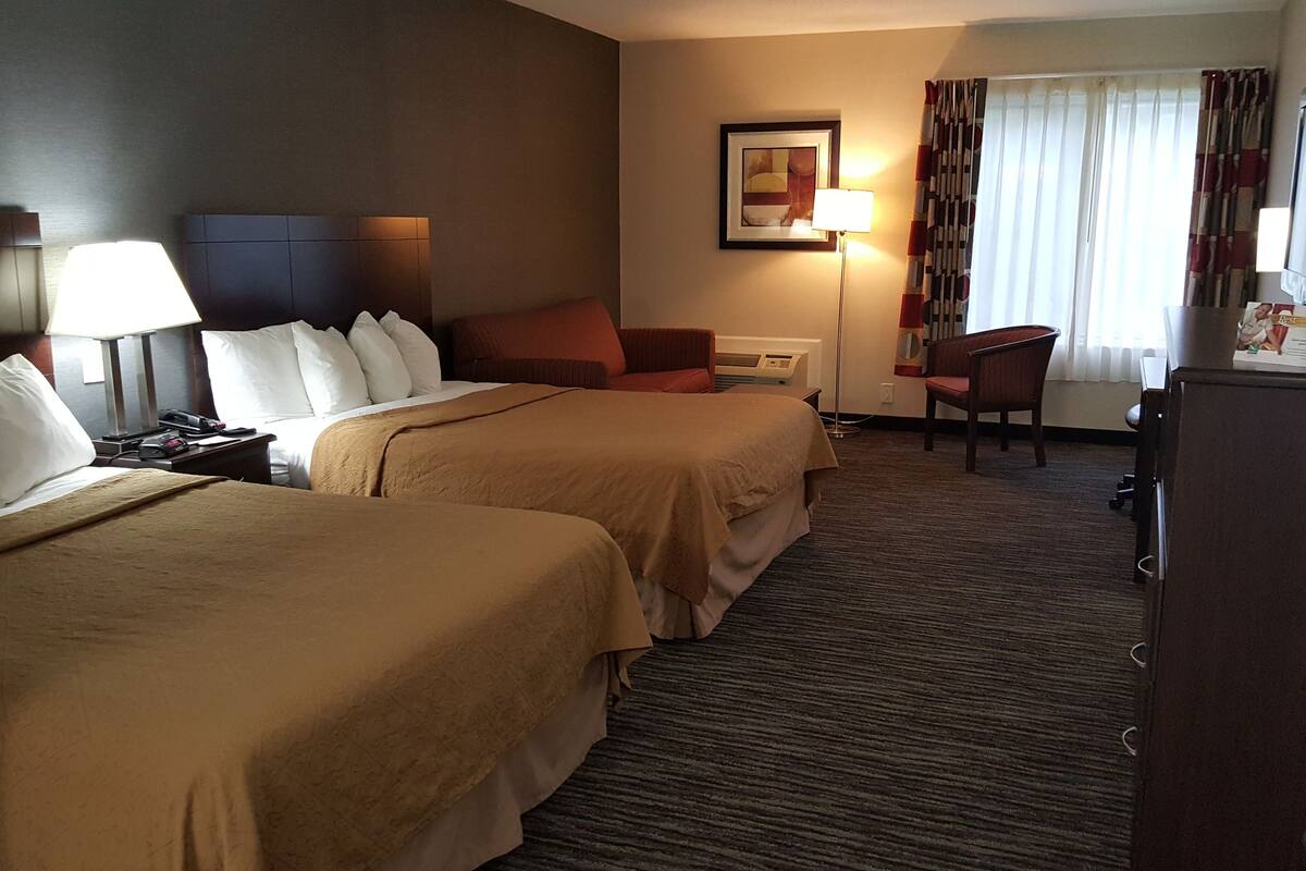A hotel room with two beds, a desk, a chair, a TV, and a lamp.