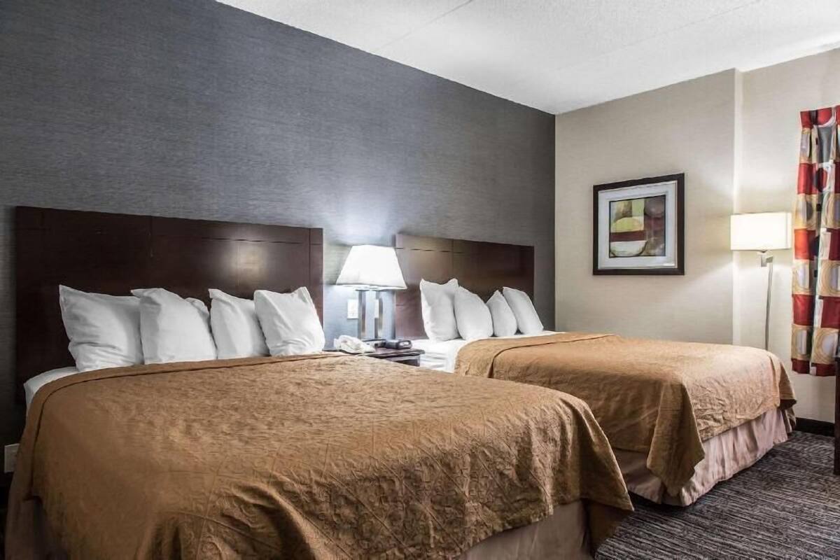 Habitación de hotel con dos camas, cabecera, lámpara, un cuadro enmarcado y cortina.