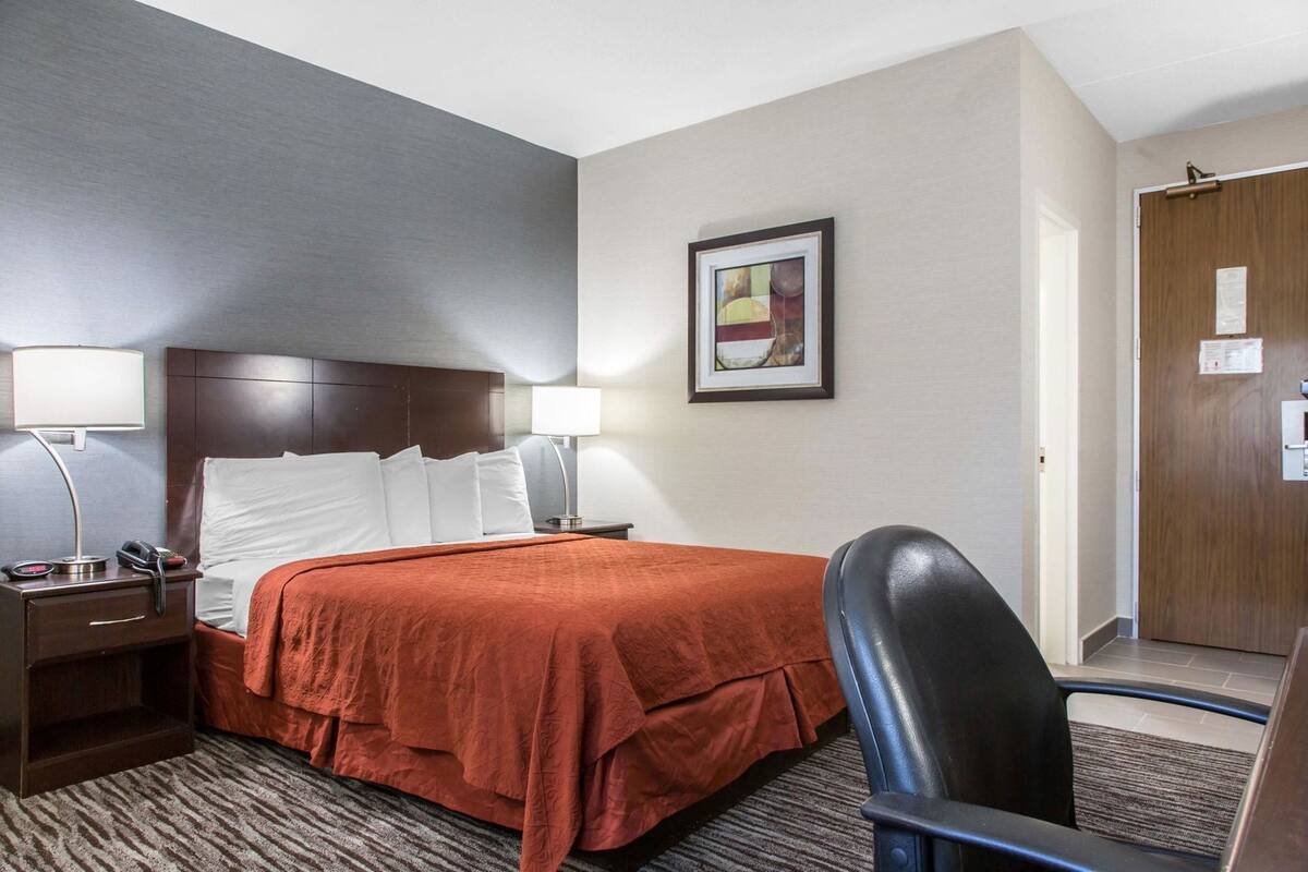 Una habitación de hotel con una cama, dos lámparas de noche, una mesita de noche con un teléfono, una silla y una puerta.