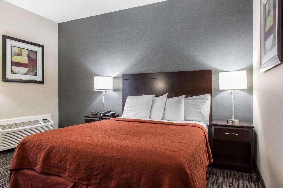 Una habitación de hotel con una cama, dos lámparas de noche, una mesita de noche, un cuadro enmarcado en la pared y un radiador.