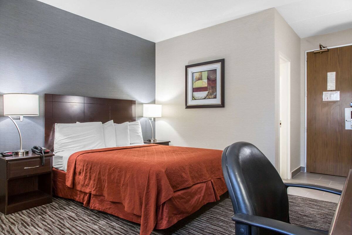 A hotel room with a bed, two bedside lamps, a desk with a chair, a framed picture on the wall, and a wooden door.