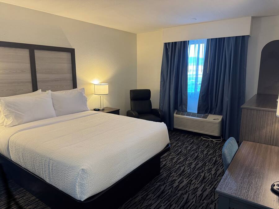 Una habitación de hotel con una cama grande, un escritorio, una silla, una lámpara y una ventana con cortinas.