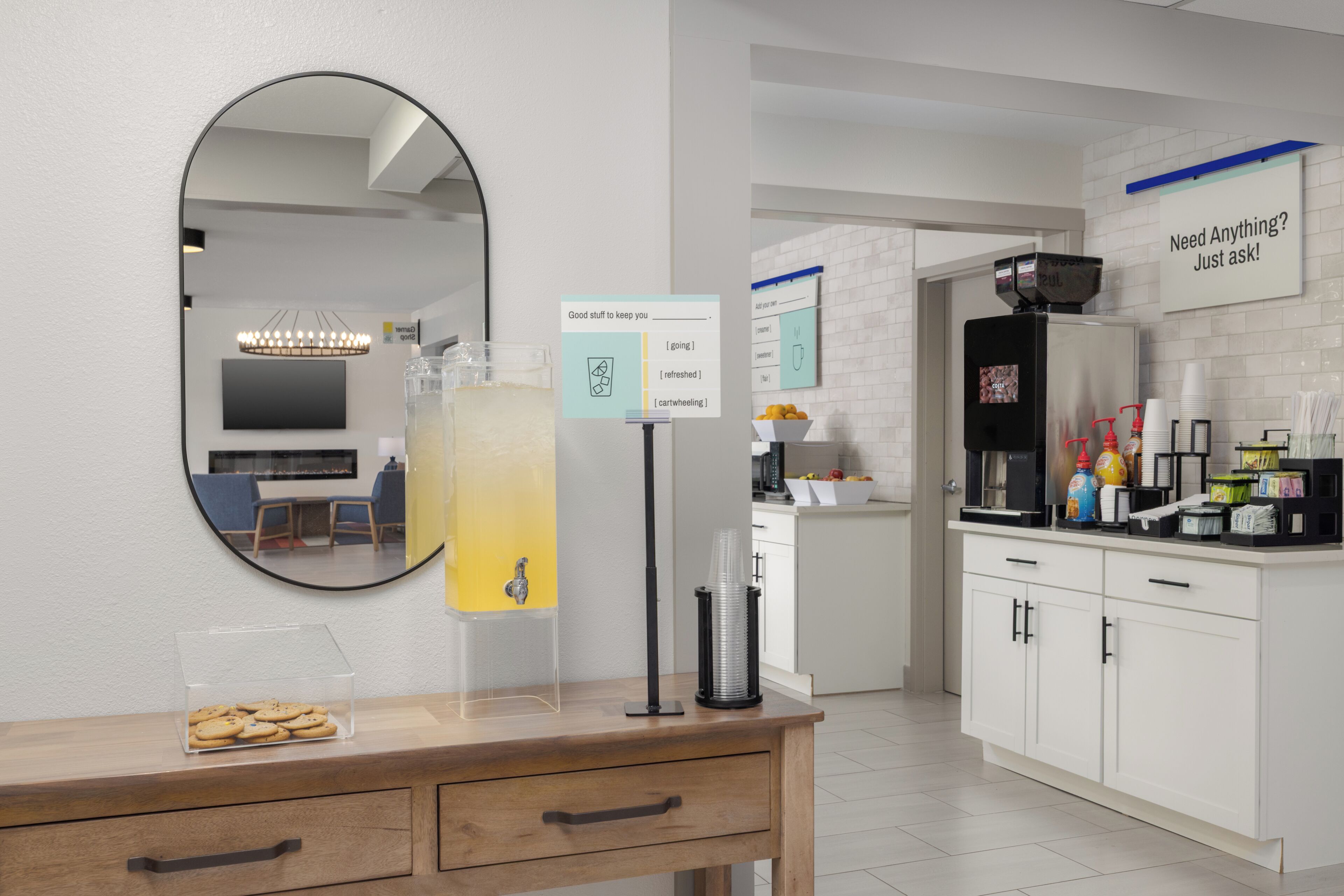 A modern kitchen with a coffee machine, a sign offering refreshment options, and a counter with a glass container of cookies.