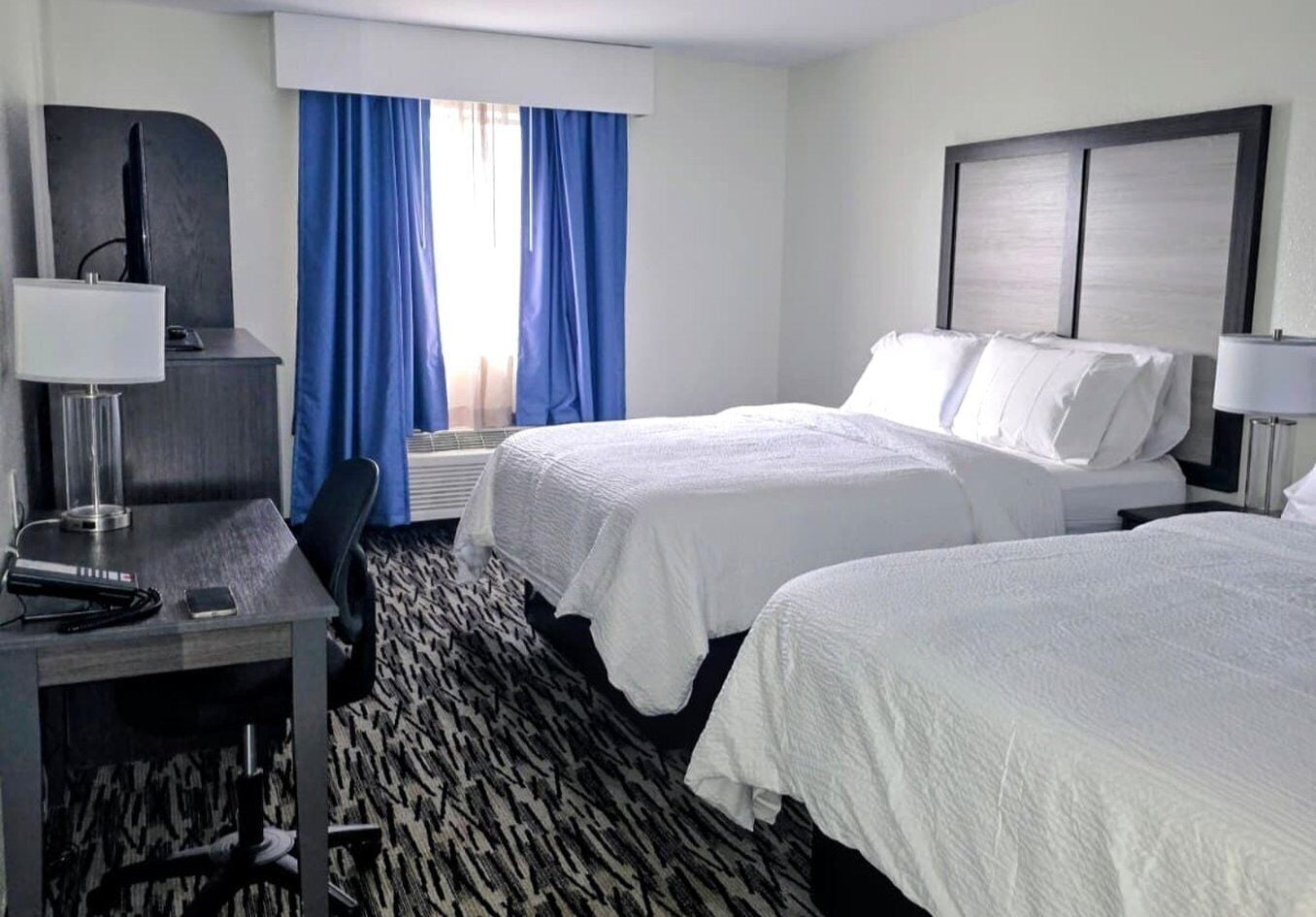A hotel room with two beds, a desk with a lamp, a chair, and a large window with blue curtains.