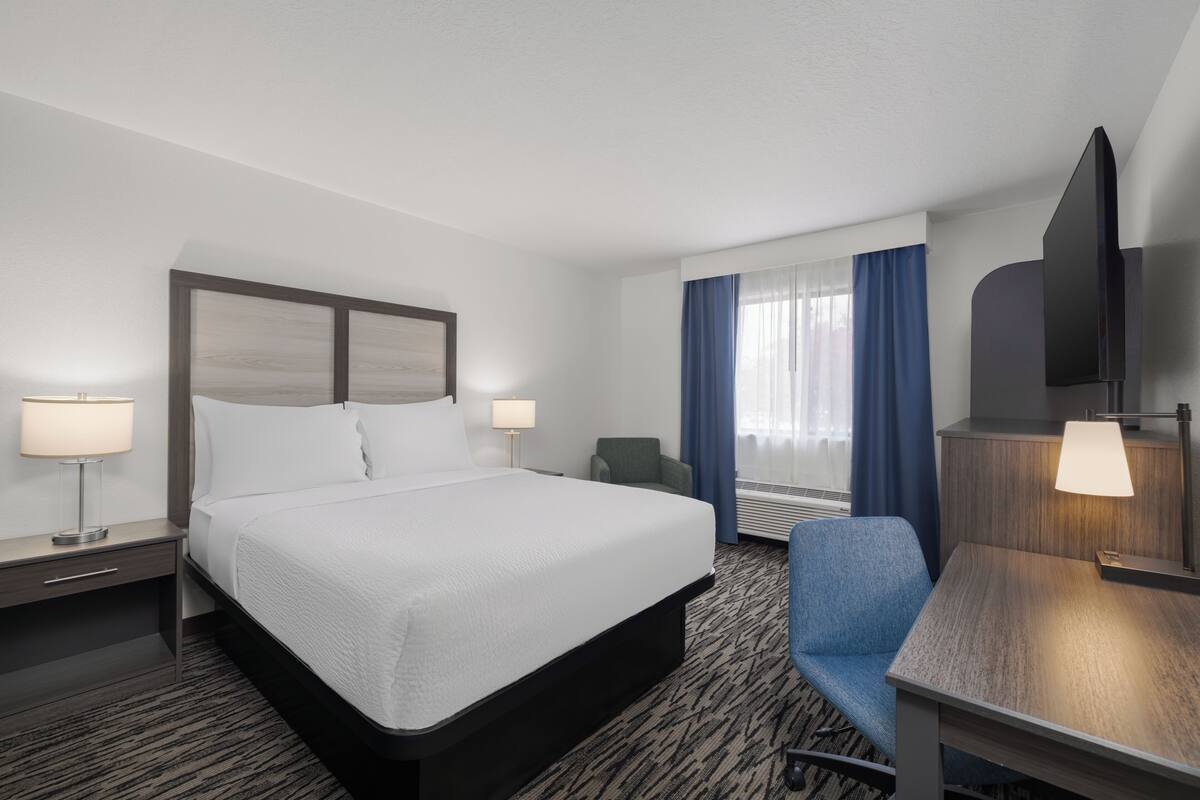 A hotel room with a bed, two bedside tables with lamps, a desk with a chair, and a large window with curtains.