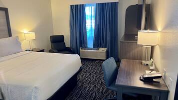 Une chambre d’hôtel avec un grand lit, un bureau avec une lampe, une chaise, une lampe de bureau, un téléphone et une fenêtre avec des rideaux.