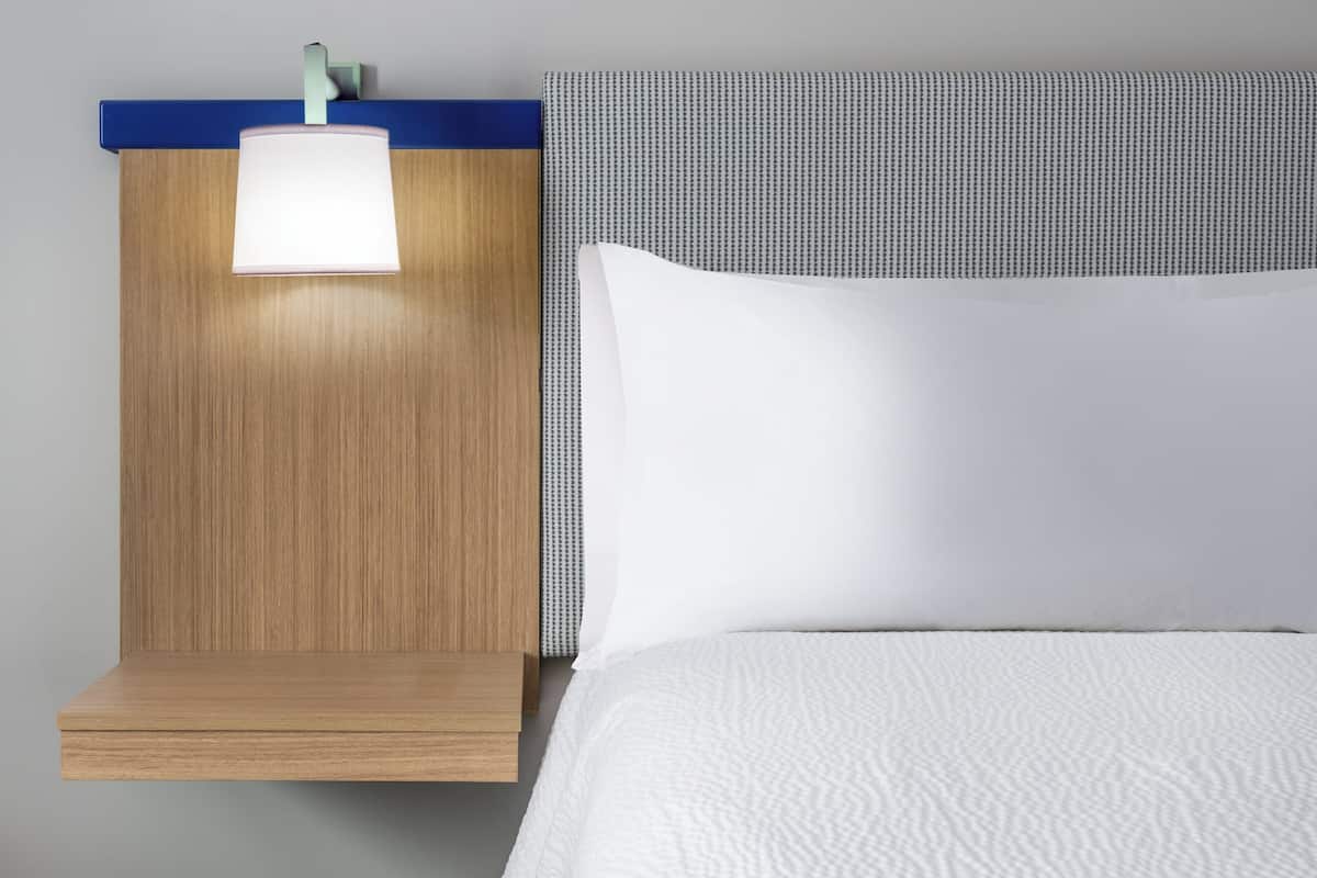 A neatly made bed with white bedding, a wooden nightstand with a lamp, and a headboard with a blue and gray pattern.