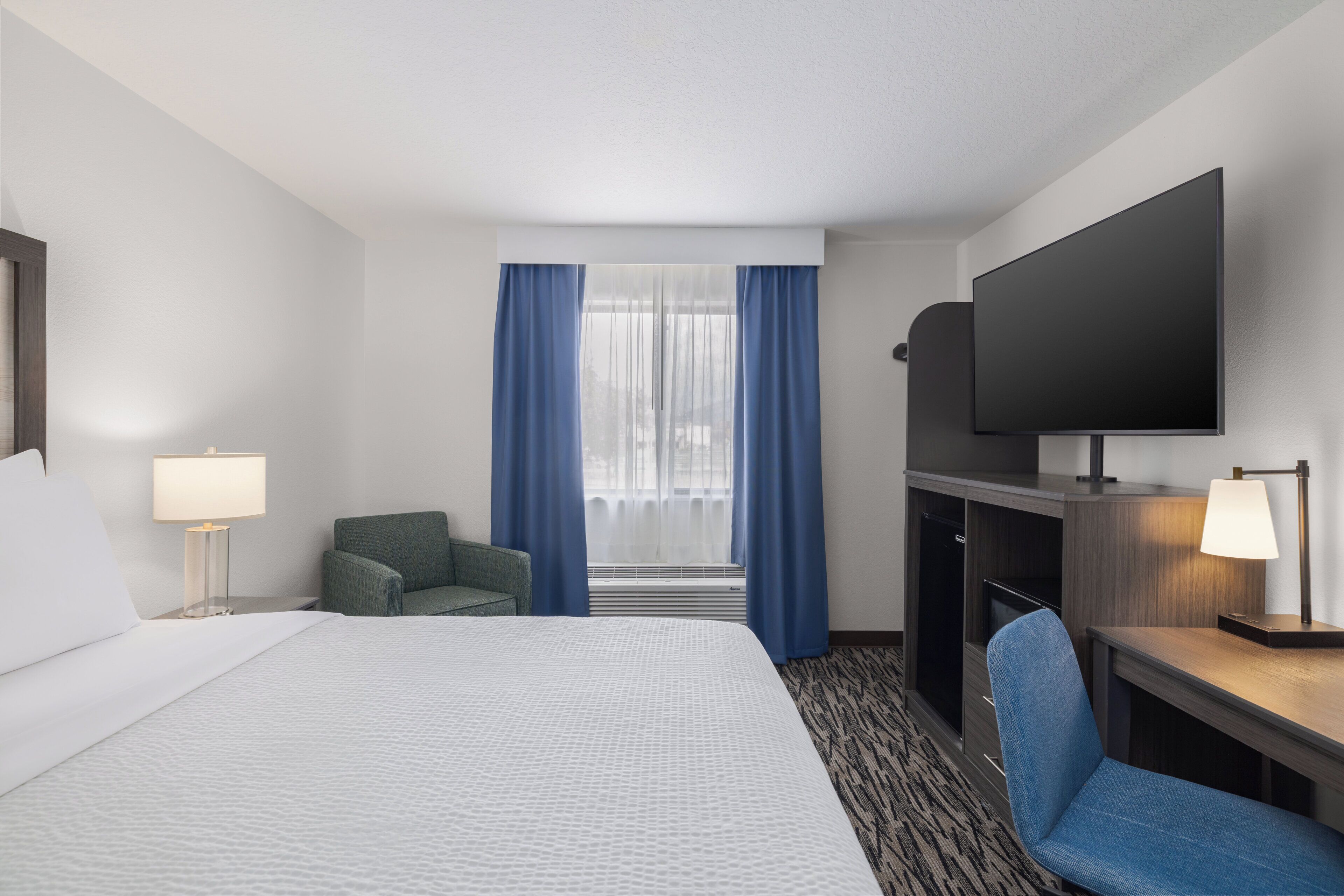 A hotel room with a bed, a desk, a chair, a TV, and a window with curtains.