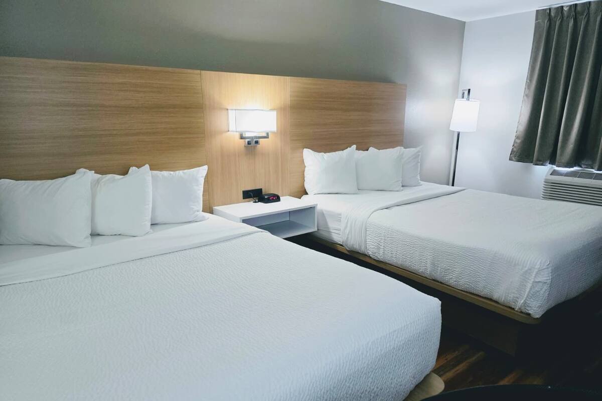 A hotel room with two beds, a nightstand, a lamp, and a window with curtains.