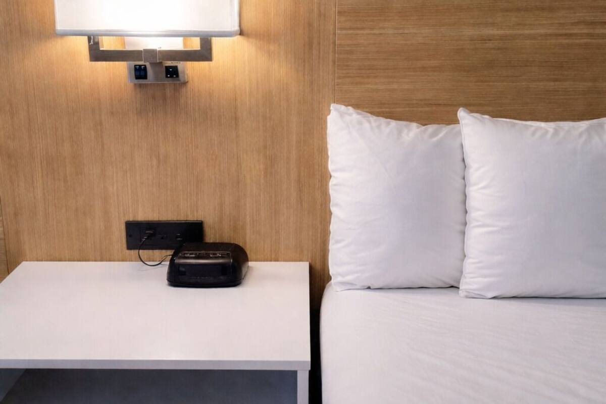 A hotel room with a wooden headboard, a bedside table with a lamp, and a bed with white bedding.