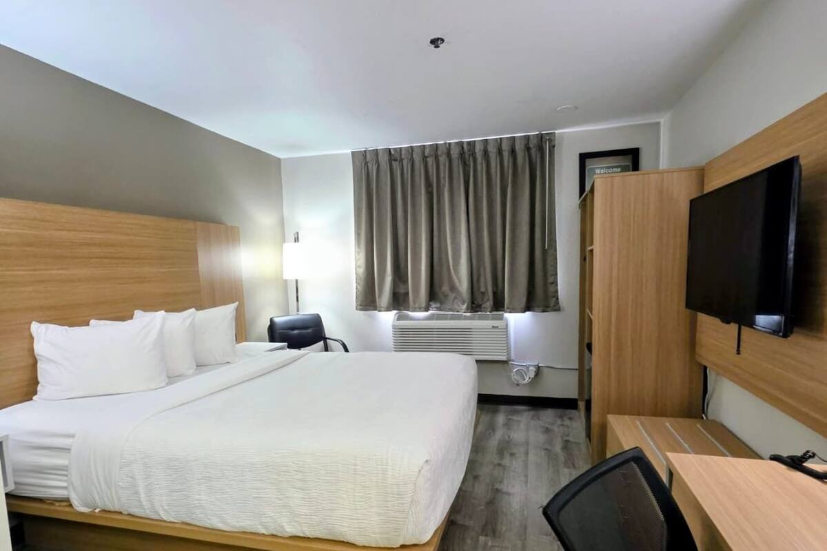 A hotel room with a bed, desk, chair, TV, and air conditioning unit.
