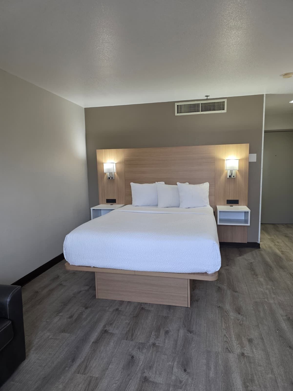 Une chambre d’hôtel avec un grand lit, deux tables de chevet avec des lampes, une tête de lit en bois et une grille d’aération au plafond.