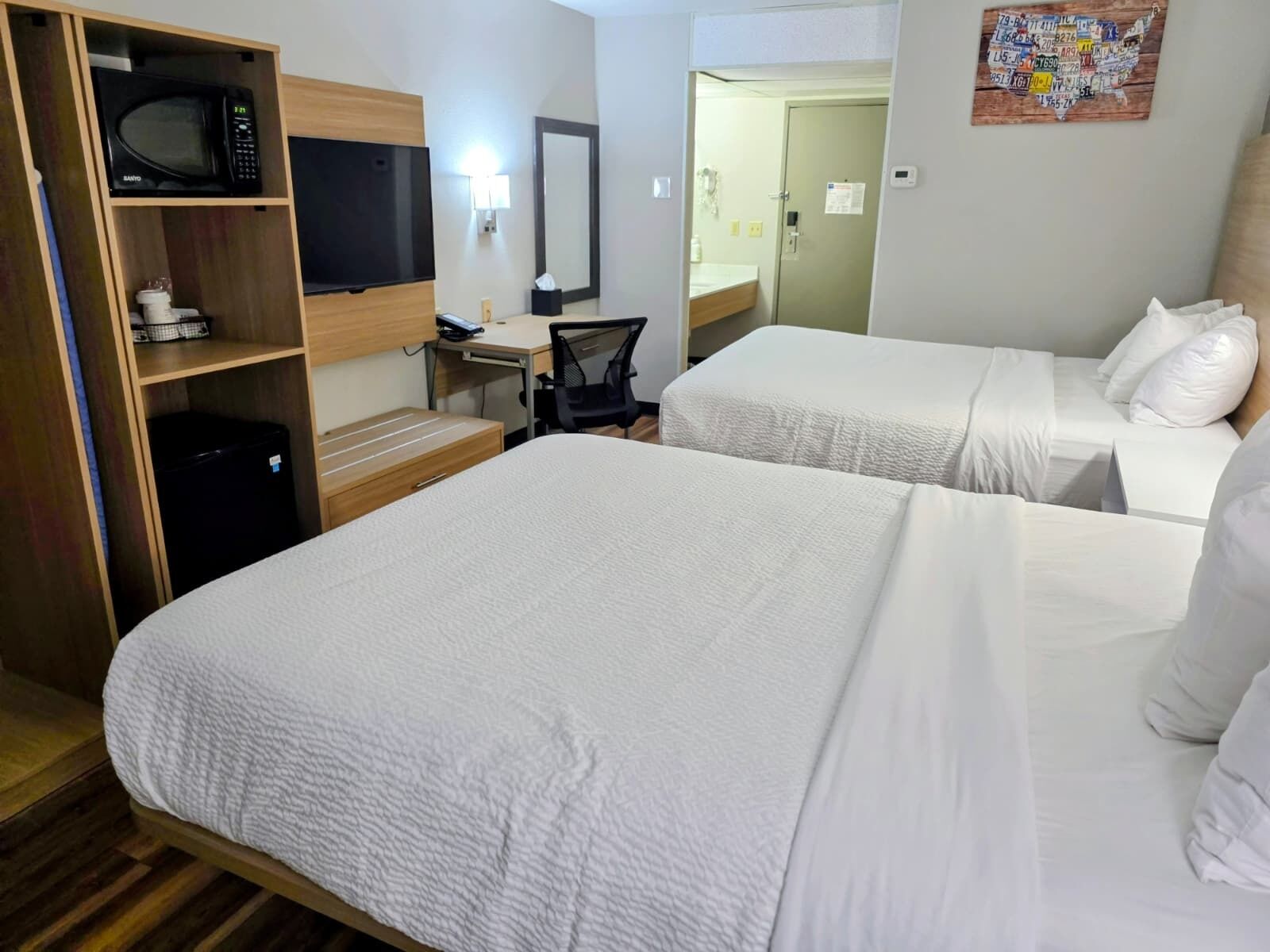 A hotel room with two beds, a desk, a chair, a microwave, a TV, and a bathroom visible through an open door.