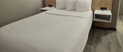 Una cama bien hecha con edredón blanco, dos mesitas de noche con lámparas y un cabecero de madera.