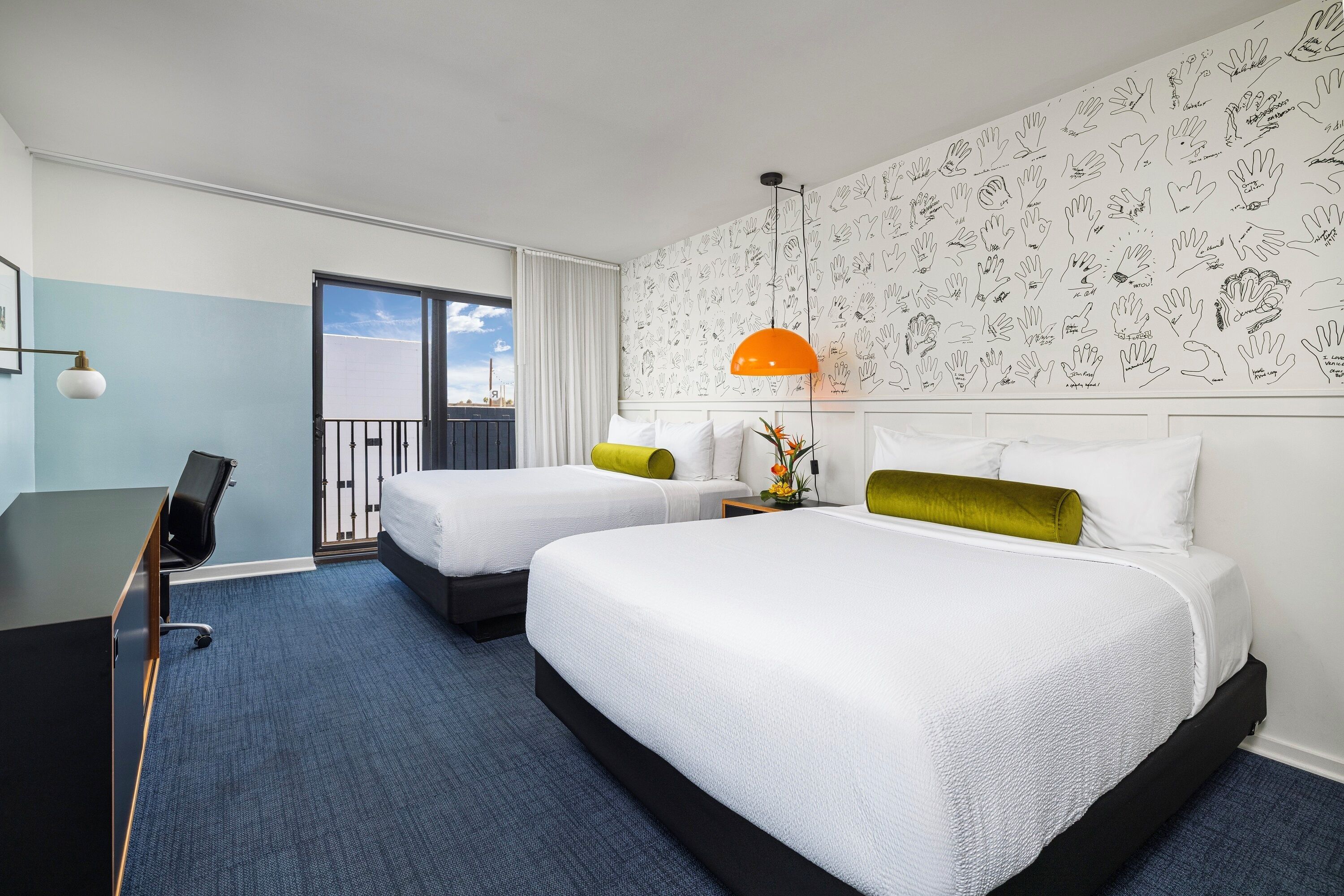 Une chambre d’hôtel avec deux lits, un bureau, une chaise, un balcon et un mur orné d’illustrations dessinées à la main.
