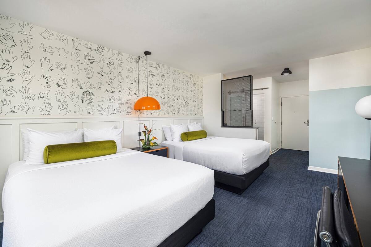 A hotel room with two beds, a desk, a chair, a window, and a patterned wall.