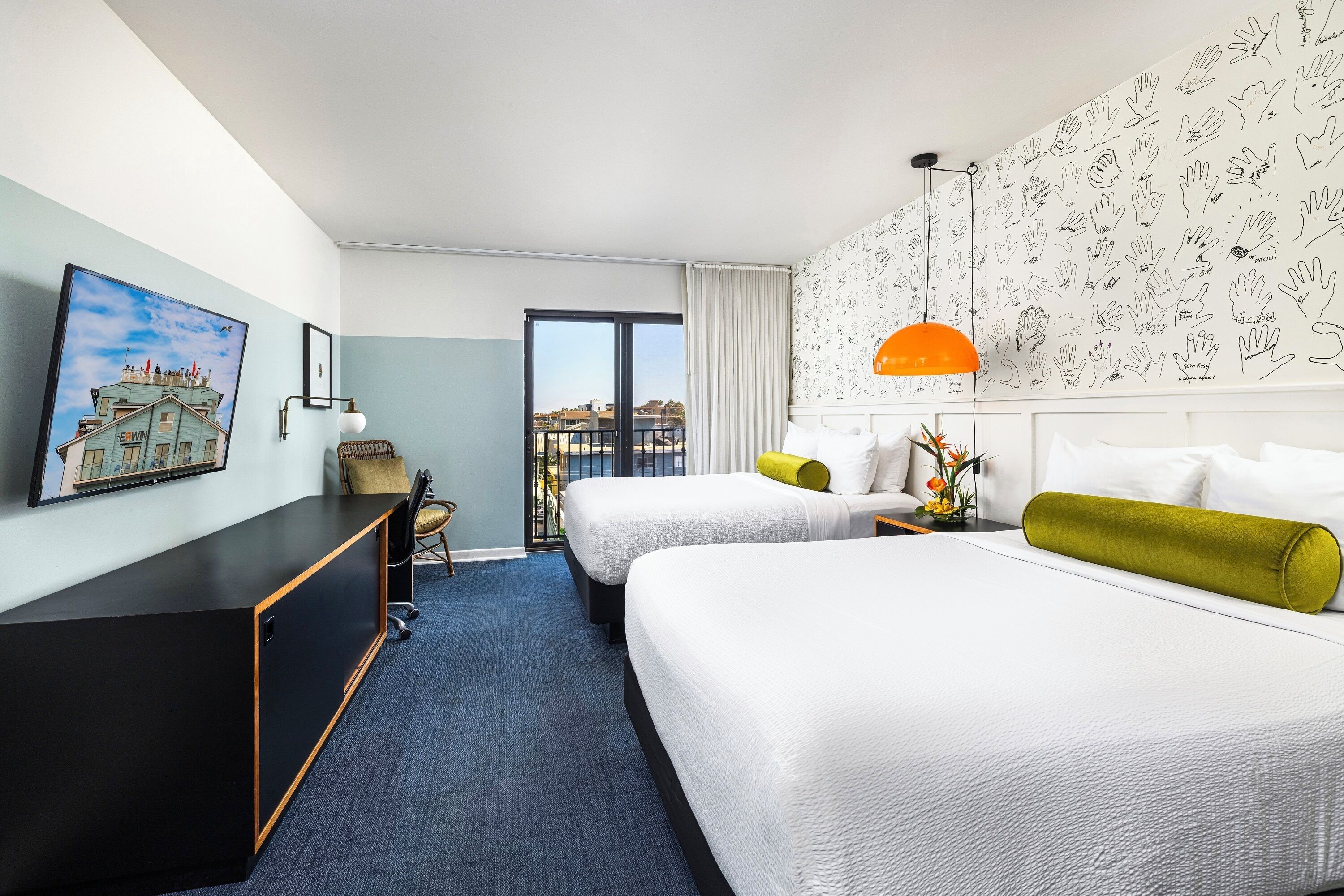 A hotel room with two beds, a desk, a chair, a window with a view, and a wall with hand-drawn illustrations.