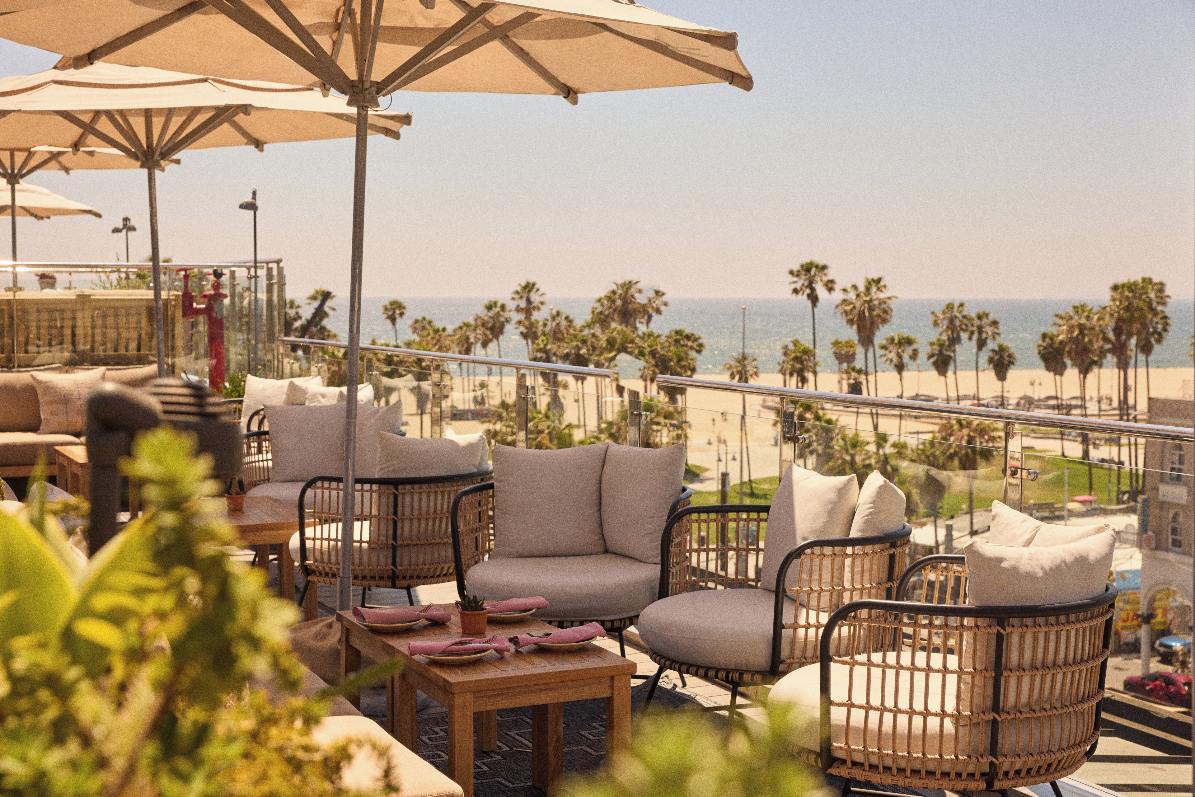 A terrace with wicker furniture, a table set for dining, and a view of the beach and palm trees.