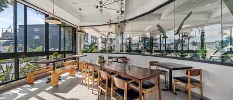 A modern dining area with wooden tables and chairs, large windows offering a view of the city, and a ceiling with recessed lighting.