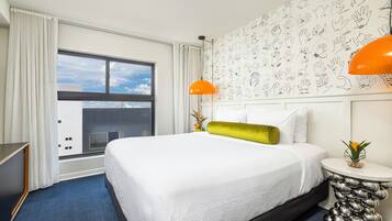 A hotel room with a large bed, a window with curtains, a wall with handprints, a small table with a plant, and a unique lamp.