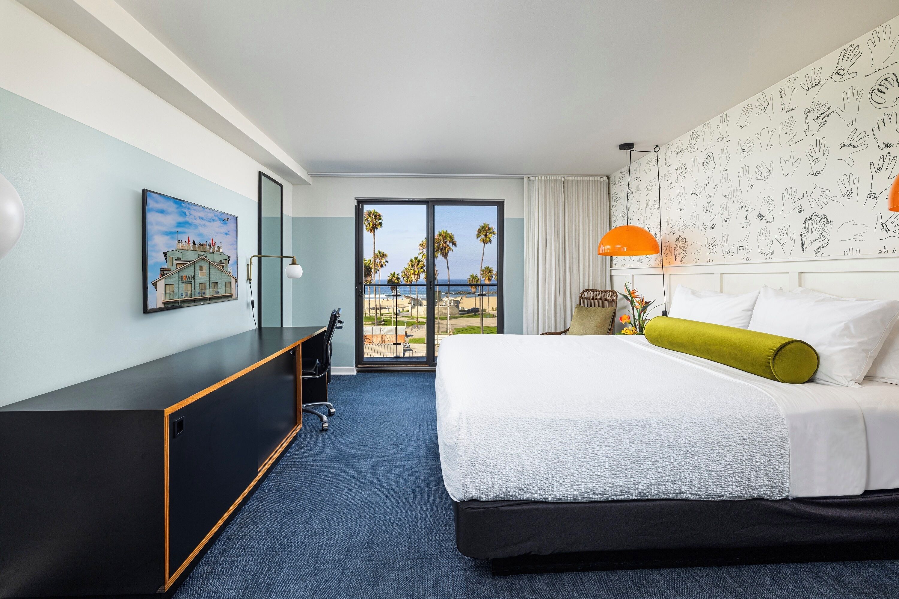 A hotel room with a bed, a desk, a chair, a large window with a view of palm trees and the ocean, and a wall with hand-drawn illustrations.