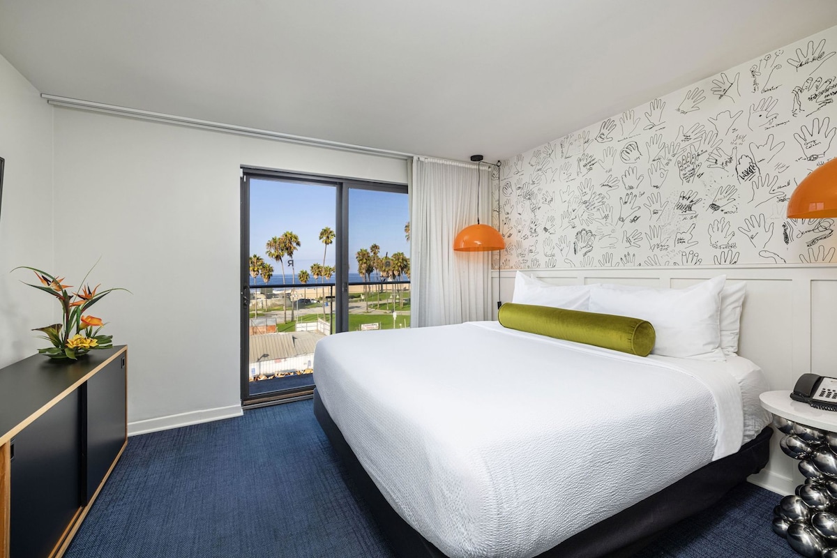 A hotel room with a large bed, a desk, a chair, a telephone, and a view of the beach.