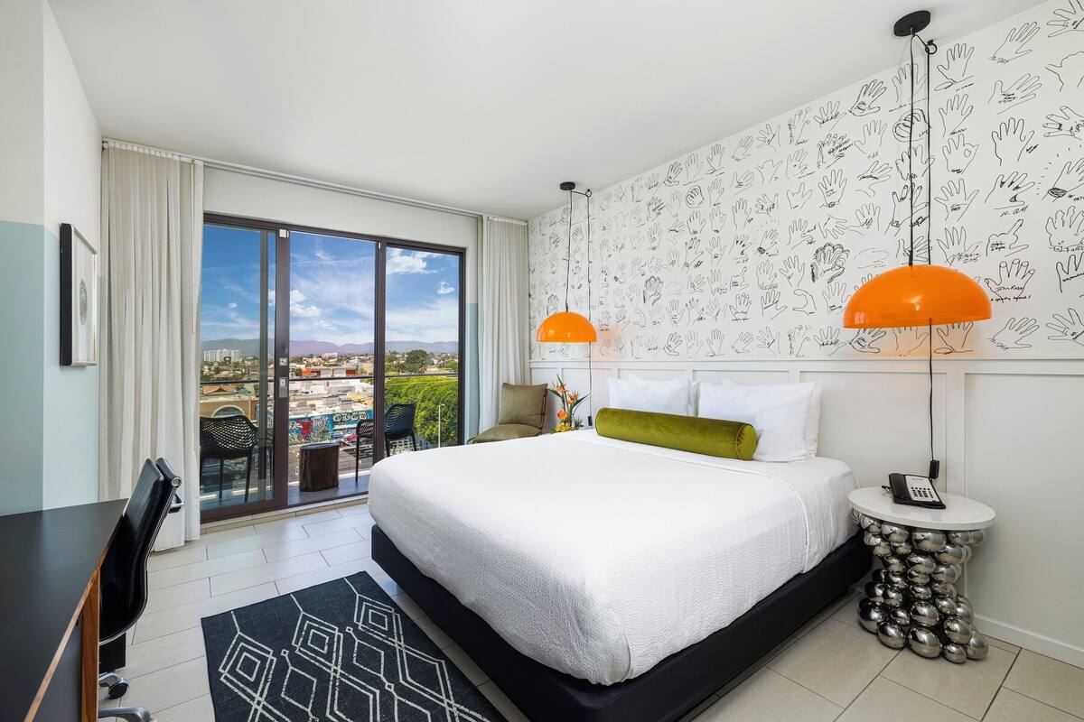A modern hotel room with a large bed, a desk, a chair, a round side table with a phone, and a view through a large window.