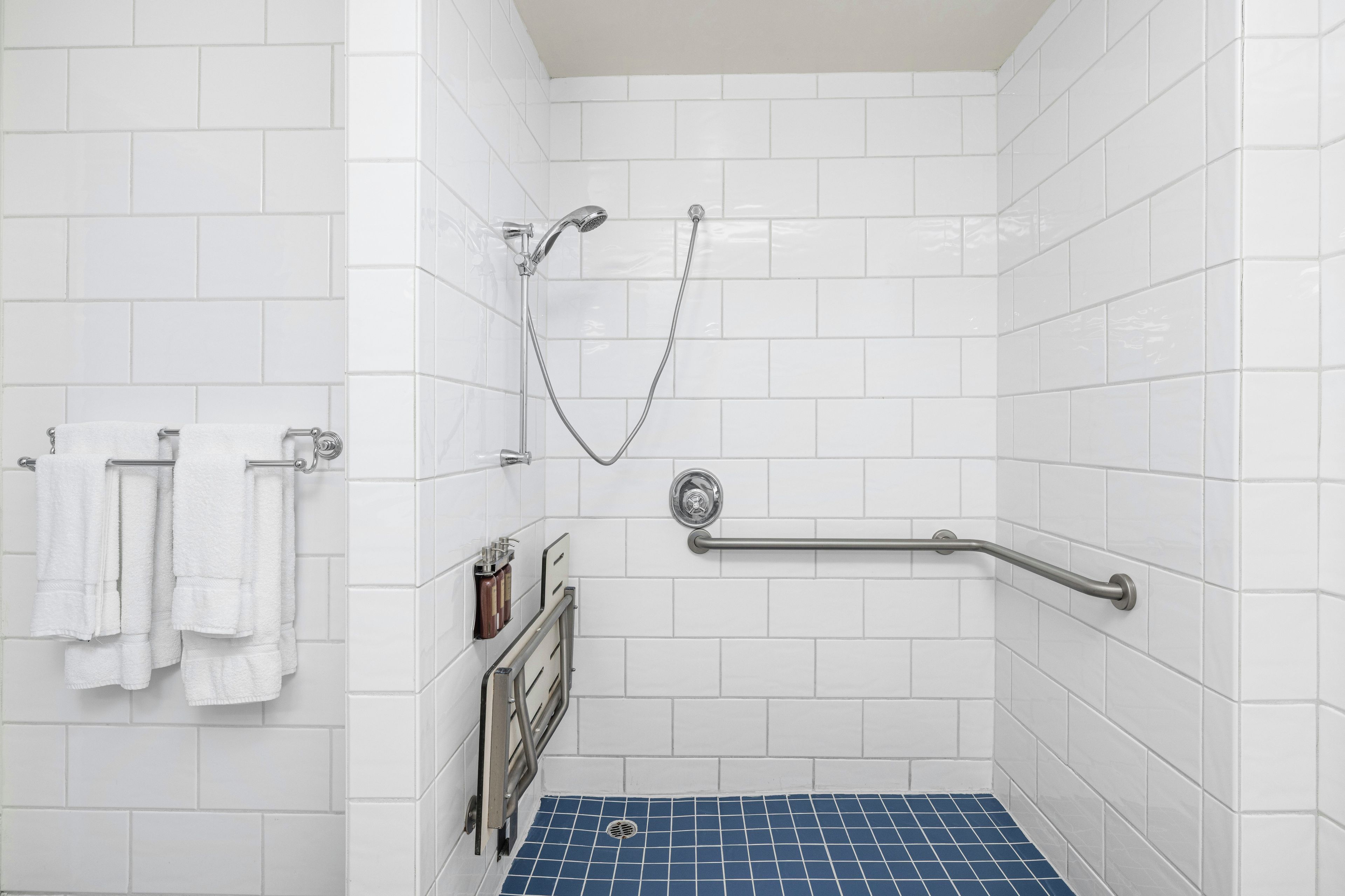 Un espace douche avec des murs recouverts de carreaux blancs, un sol carrelé de bleu, une douchette à main, une barre d’appui et un porte-serviettes avec des serviettes pliées.
