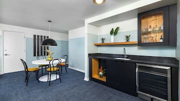 A compact kitchen with a built-in oven, a dining area with a table and chairs, and a small shelf with plants and bottles.