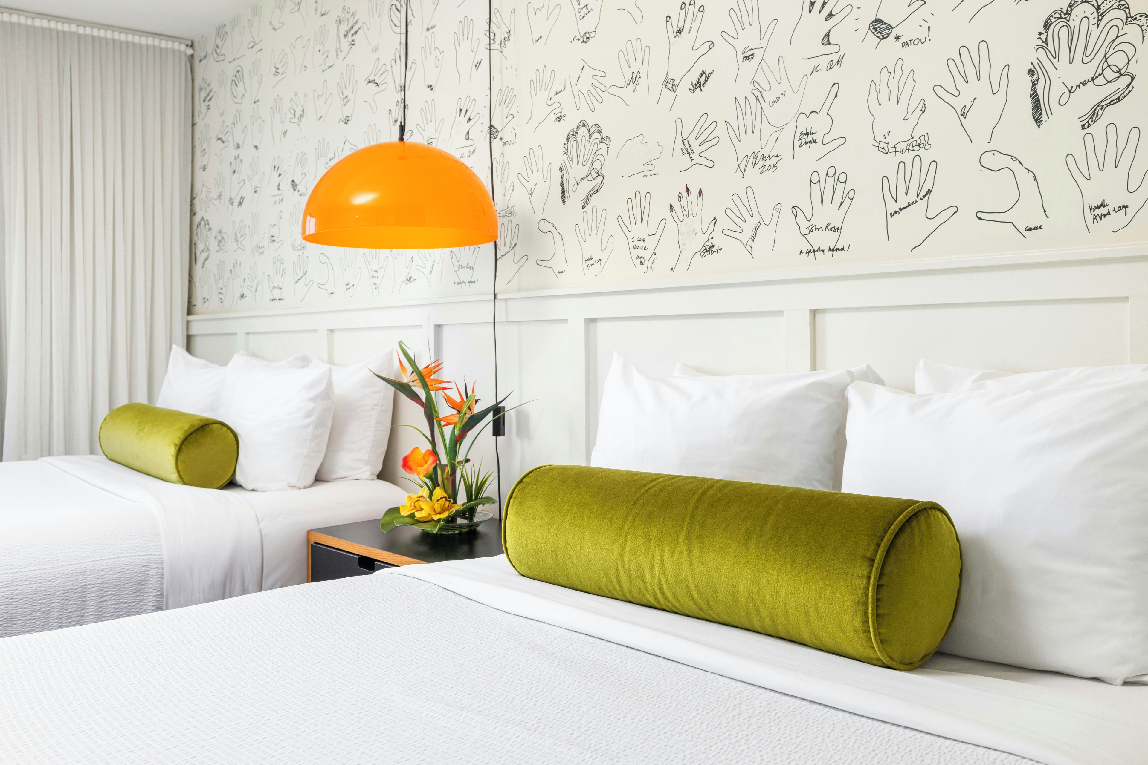 A hotel room with two beds, white bedding, green pillows, a nightstand with a vase of flowers, and a wall with hand-drawn handprints.