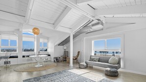 A spacious loft-style room with a high ceiling, large windows offering a view of the beach, a gray sofa with patterned cushions, and a dining area with a round table and chairs. - Hotel Erwin Venice Beach (Venice)
