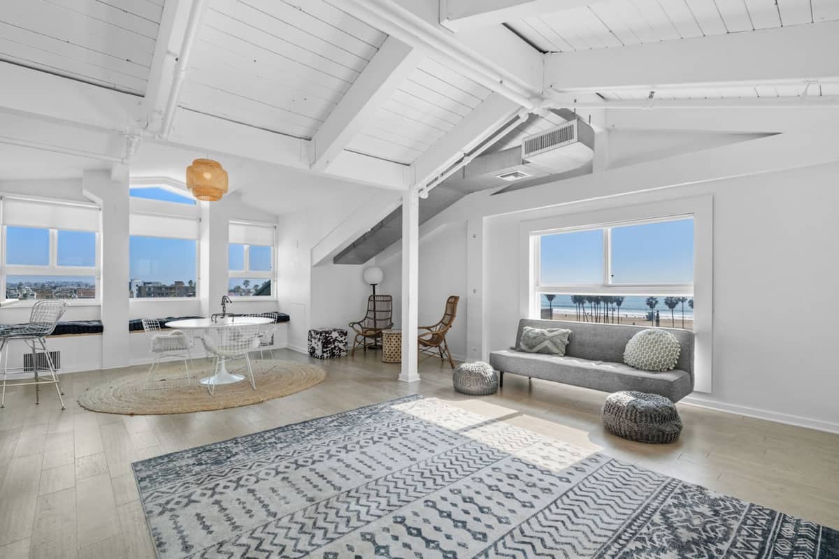 A spacious loft-style room with a high ceiling, large windows offering a view of the beach, a gray sofa with patterned cushions, and a dining area with a round table and chairs.