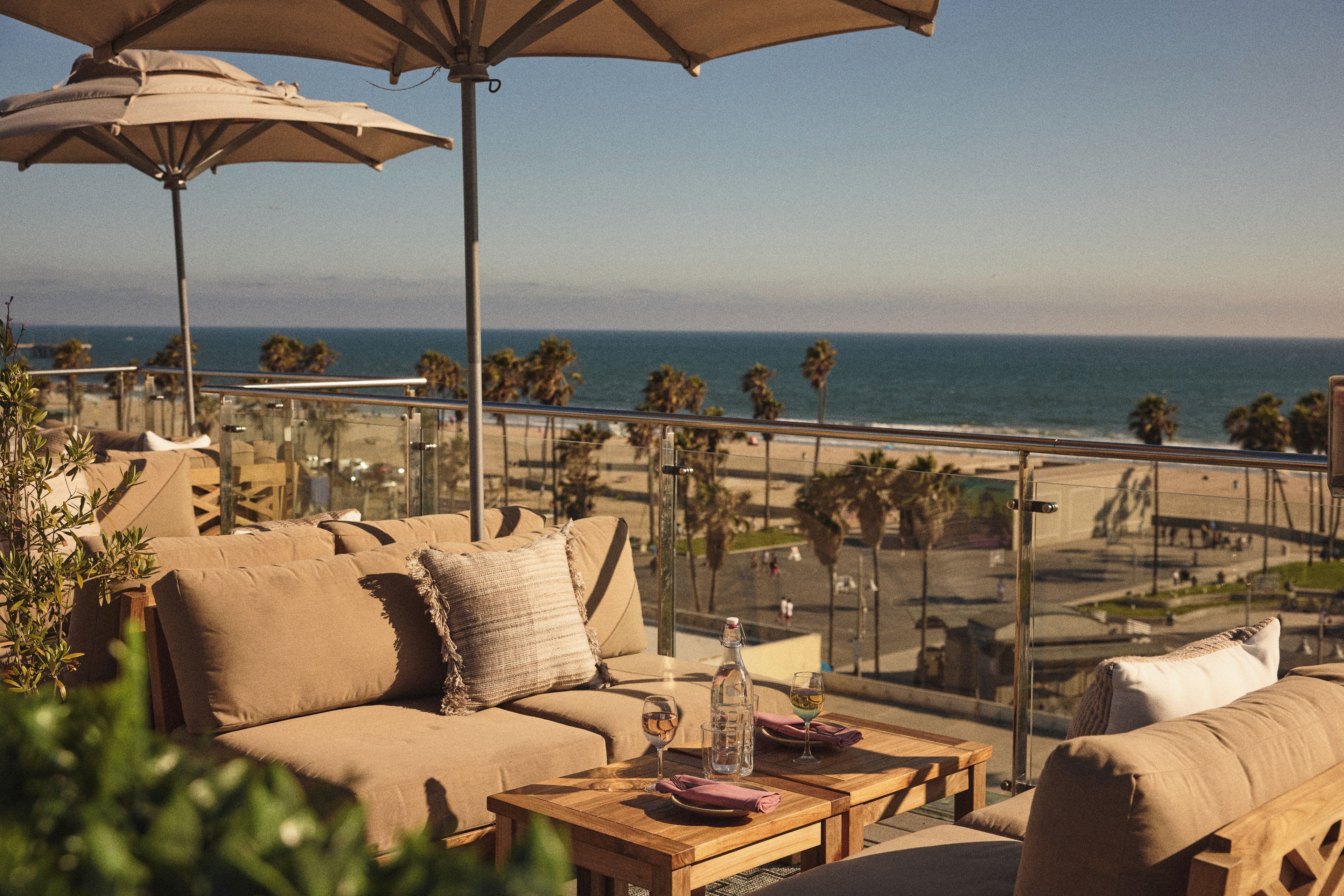A balcony with a sofa, a table, and umbrellas overlooking a beach with palm trees and the ocean.