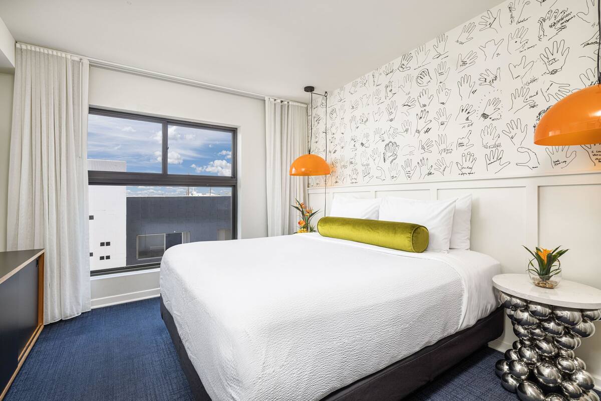 A hotel room with a large bed, a window with curtains, a wall with hand-drawn patterns, and a unique metallic side table.