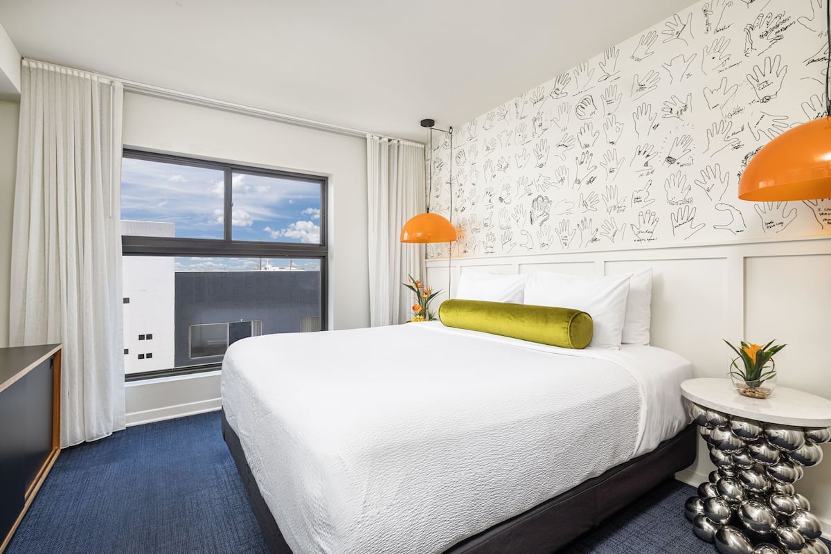 A hotel room with a large bed, a window with curtains, a wall with hand-drawn patterns, and a unique metallic side table.