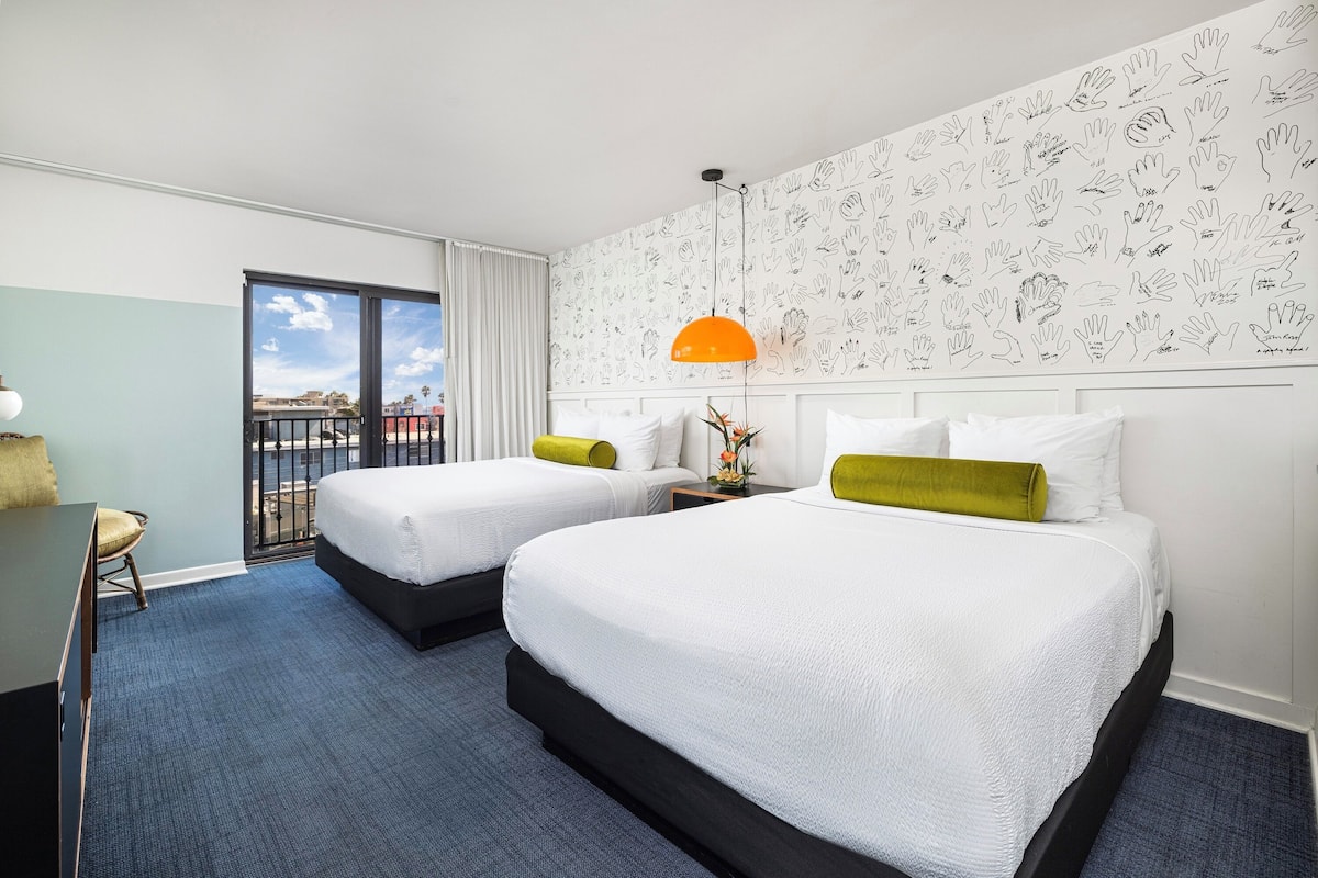 A hotel room with two beds, a desk, a chair, a balcony, and a wall with hand-drawn patterns.