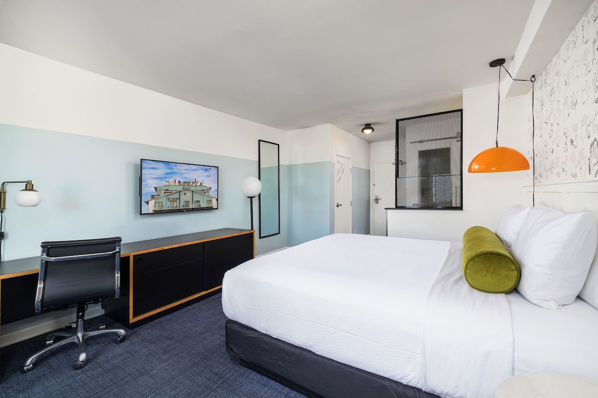 A hotel room with a bed, desk, chair, and a painting on the wall.