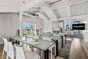 A modern hotel room with a large window overlooking a beach, a flat-screen TV mounted on the wall, and a dining area with tables set for guests. - Hotel Erwin Venice Beach (Venice)